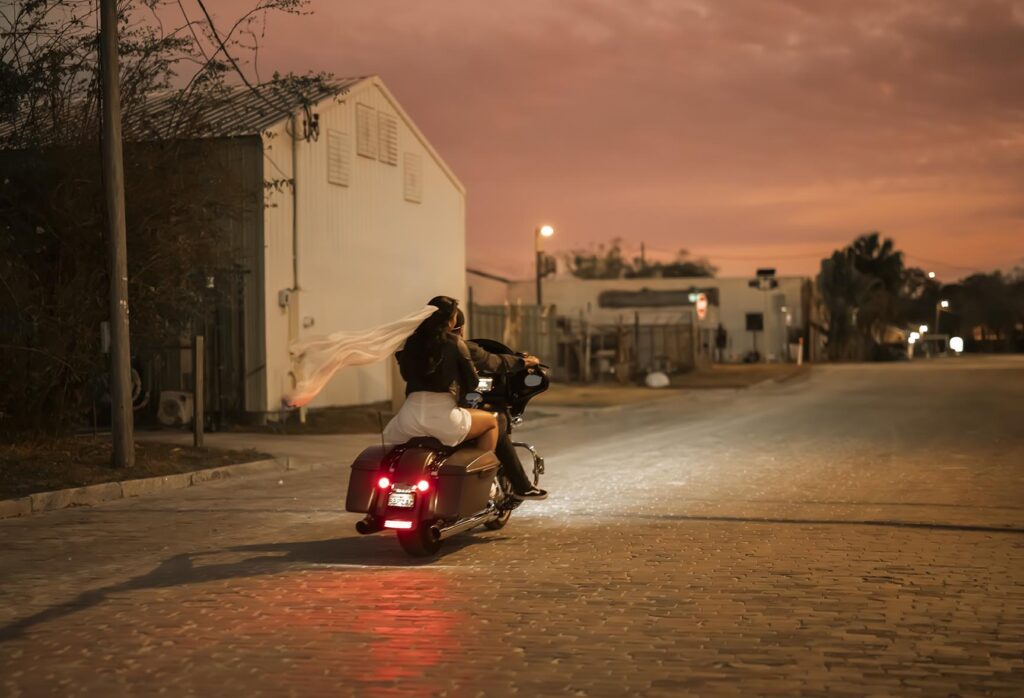 A woman with a flowing white scarf rides a motorcycle down a quiet street at sunset, with buildings and trees lining the road.