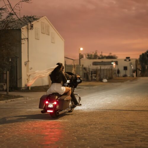 A woman with a flowing white scarf rides a motorcycle down a quiet street at sunset, with buildings and trees lining the road.