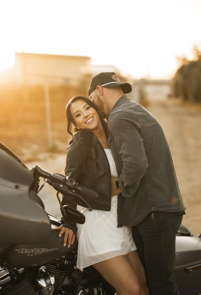 A woman sits on a Harley-Davidson motorcycle, smiling at the camera, while a man stands close beside her outdoors in warm sunlight.