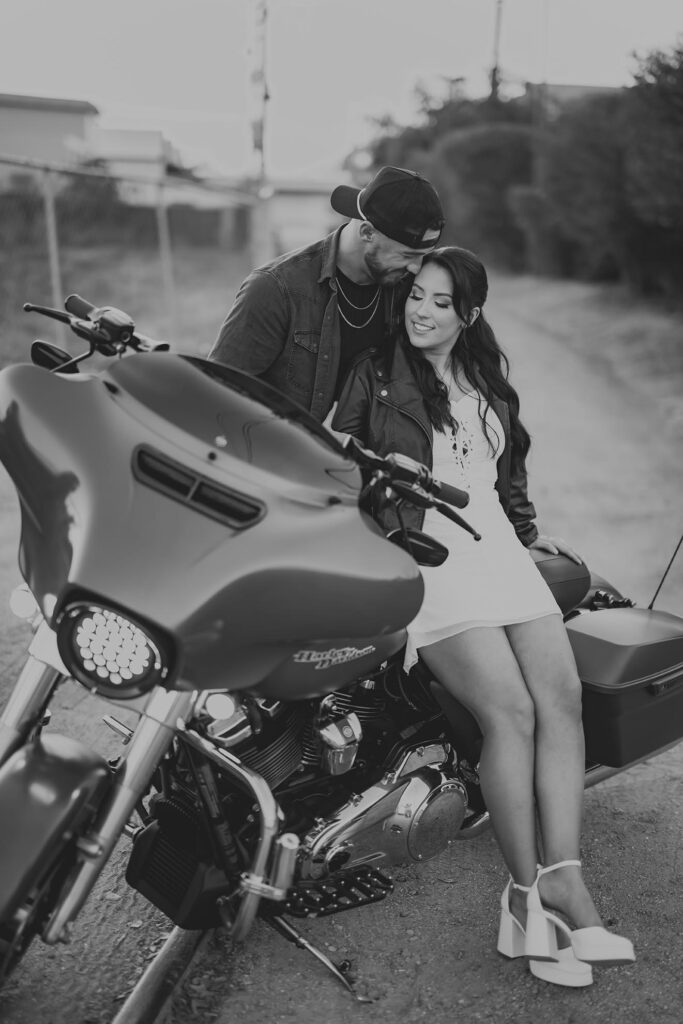 A man stands close to a seated woman as she sits on a motorcycle; both are smiling and appear relaxed on a dirt path.