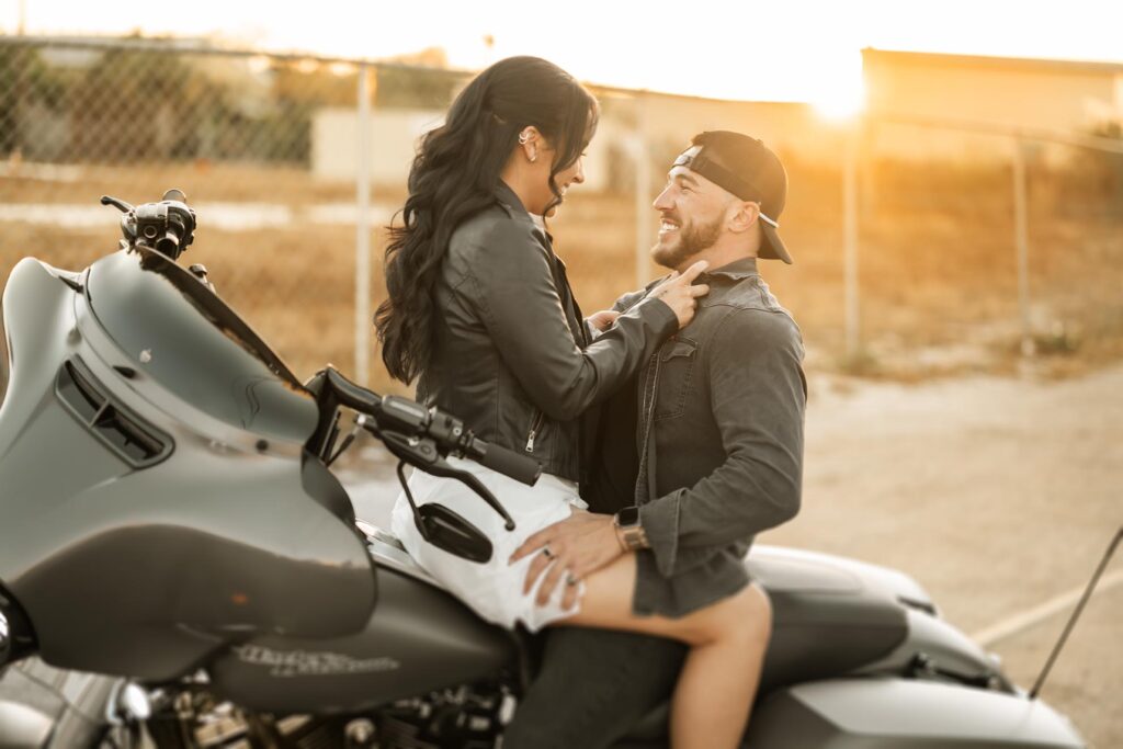 A woman sits on a motorcycle facing a smiling man, both wearing jackets. The man holds her waist, and the scene is outdoors at sunset near a fence.