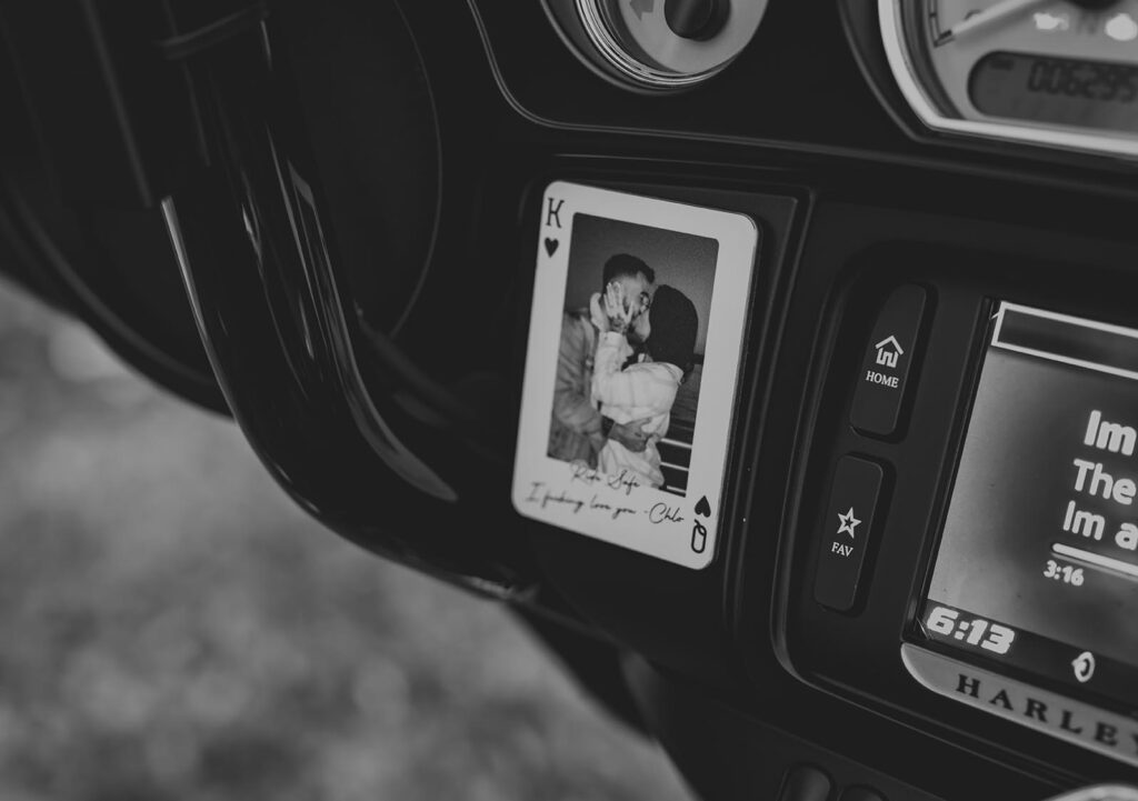 Black and white photo of a King of Hearts card with a couple kissing, placed on a Harley-Davidson motorcycle dashboard near the radio display.